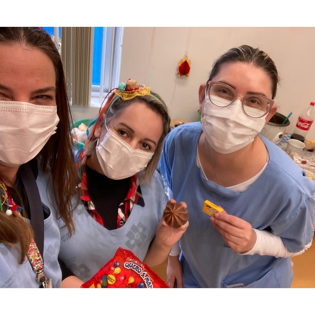 Três mulheres profissionais da saúde seguram doces da festa junina e posam pra foto com uniforme e máscara