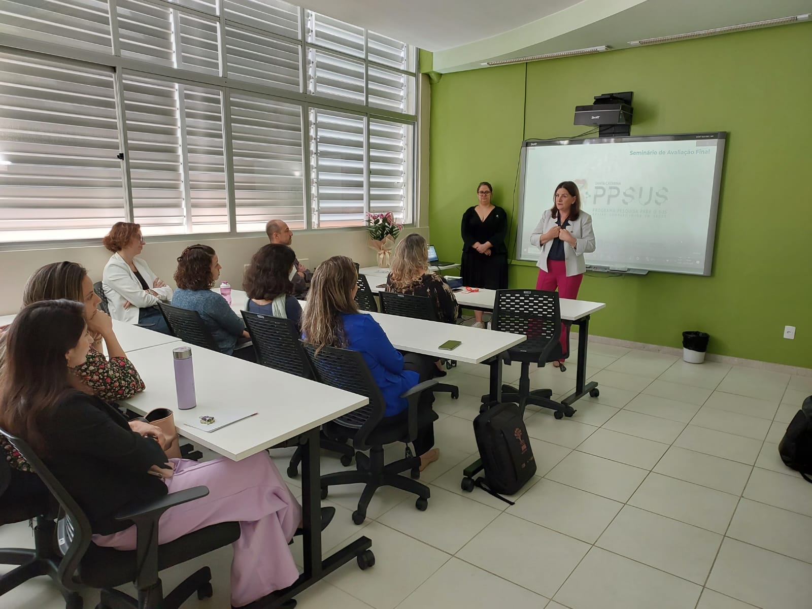 A imagem mostra uma sala de aula ou treinamento com paredes verdes e janelas amplas com persianas metálicas. Cerca de dez pessoas estão presentes, sentadas em cadeiras pretas com mesas brancas, voltadas para a frente da sala. Duas mulheres estão em pé, conduzindo a apresentação: uma delas veste calça rosa e blazer claro, enquanto a outra usa um vestido preto.  No fundo, há um quadro interativo ou tela de projeção, exibindo a frase "Seminário de Avaliação Final – PPSUS", indicando que o evento é voltado para a avaliação de um programa ligado à saúde pública e pesquisa. O ambiente tem clima formal e voltado para capacitação ou encerramento de um projeto.