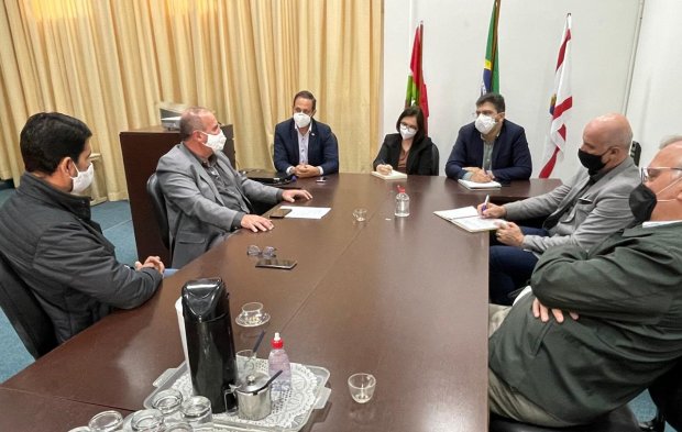 Reunião formal com sete pessoas sentadas ao redor de uma mesa de madeira, todos usando máscaras faciais. Na mesa há copos, garrafa térmica, álcool em gel e documentos. Ao fundo, há três bandeiras: a de Santa Catarina, do Brasil e uma bandeira institucional. As cortinas amarelas cobrem a parede ao fundo, e o ambiente transmite um clima sério e institucional.