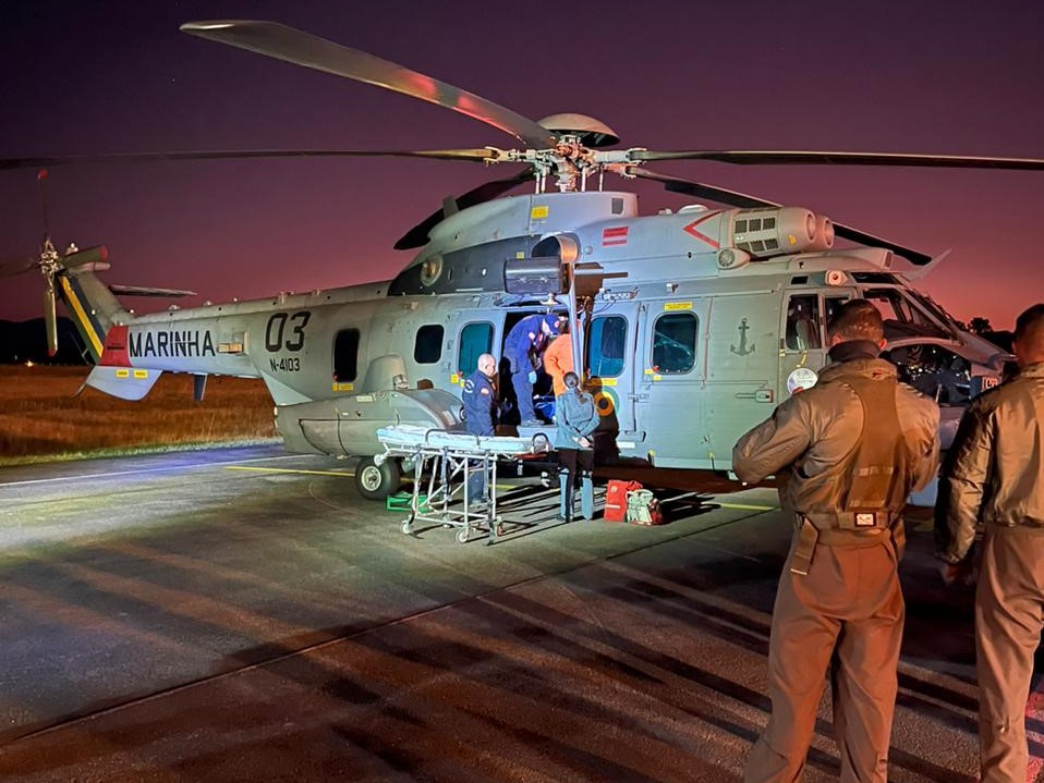 Equipes de resgate realizam atendimento médico em um helicóptero da Marinha do Brasil durante a noite. A aeronave modelo UH-15 Super Cougar, com número 03, está estacionada em uma pista iluminada artificialmente. Profissionais uniformizados operam equipamentos médicos e preparam uma maca para transporte de paciente. O céu ao fundo apresenta tons de roxo e laranja do entardecer.