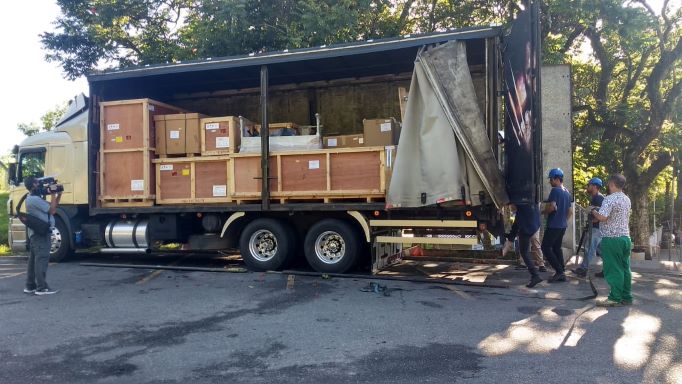 Caminhão baú com a lateral aberta mostrando diversas caixas e engradados de madeira empilhados em seu interior, indicando transporte de equipamentos ou materiais. À direita, trabalhadores com capacetes azuis e roupas de segurança acompanham o descarregamento. À esquerda, uma pessoa com câmera profissional registra a cena. A ação ocorre em uma área externa com árvores ao fundo.