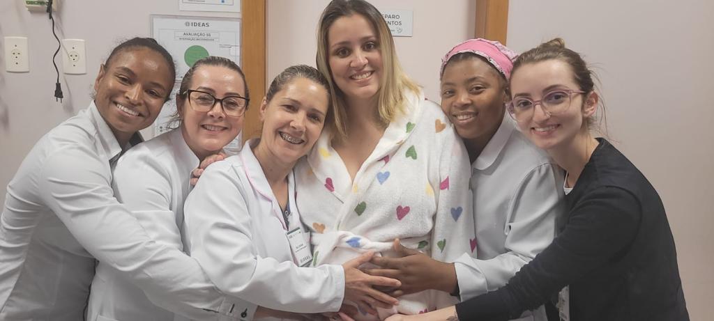 Grupo de seis mulheres sorrindo e abraçando uma gestante que veste um roupão branco com estampas coloridas de corações. As mulheres ao redor usam jalecos brancos e demonstram carinho e acolhimento. A foto transmite um momento de afeto, cuidado e apoio à futura mãe.