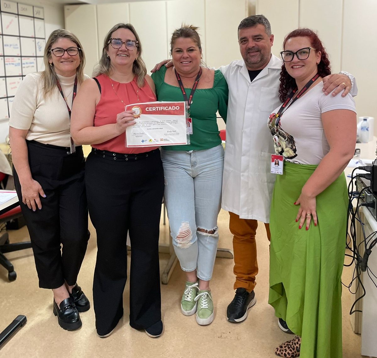 A imagem mostra um grupo de cinco pessoas sorrindo e posando para a foto em um ambiente interno, possivelmente de trabalho ou institucional. A mulher ao centro segura um certificado e veste blusa coral e calça preta; ela está acompanhada por duas mulheres à esquerda, uma usando blusa bege e outra verde, e à direita por um homem de jaleco branco e uma mulher com saia verde e camiseta estampada.  As pessoas estão próximas e abraçadas, transmitindo clima de celebração e reconhecimento, provavelmente relacionado a uma conquista ou homenagem. Ao fundo, há mesas, cadeiras e um painel com anotações, reforçando o cenário de um espaço profissional ou administrativo.