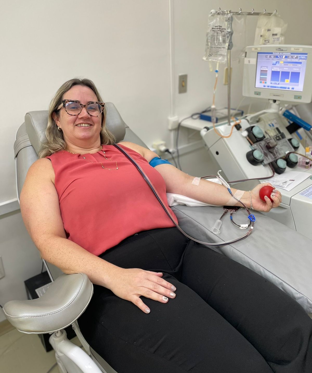 A imagem mostra uma mulher sentada em uma poltrona de hemoterapia, sorrindo, enquanto realiza um procedimento de doação de sangue ou aférese. Ela veste blusa sem mangas na cor coral e calça preta, usando óculos de armação estampada.  Seu braço direito está conectado por um tubo a um equipamento médico automatizado, visível ao fundo, que processa o sangue. Ela segura uma pequena bola vermelha na mão para auxiliar a circulação durante a coleta. Ao lado do equipamento, há bolsas e frascos pendurados com fluídos relacionados ao procedimento.  O ambiente é limpo, bem iluminado e típico de um setor hospitalar ou banco de sangue.
