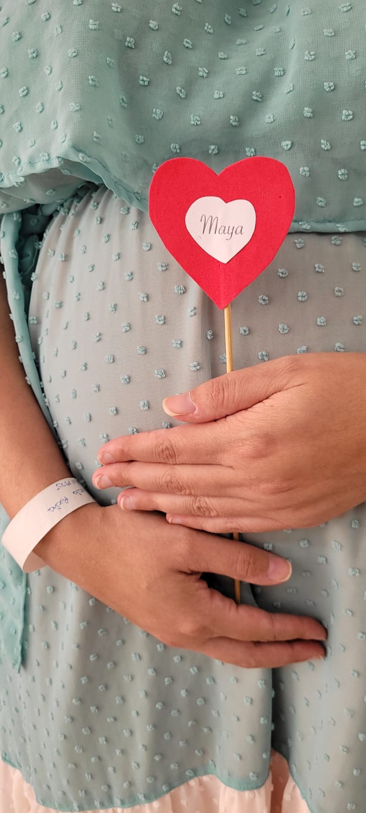 Close de uma barriga de gestante coberta por um vestido azul claro com textura de bolinhas. A futura mãe segura um pequeno enfeite em forma de coração vermelho com o nome "Maya" escrito no centro, simbolizando o nome do bebê. Ela também usa uma pulseira hospitalar no pulso esquerdo.