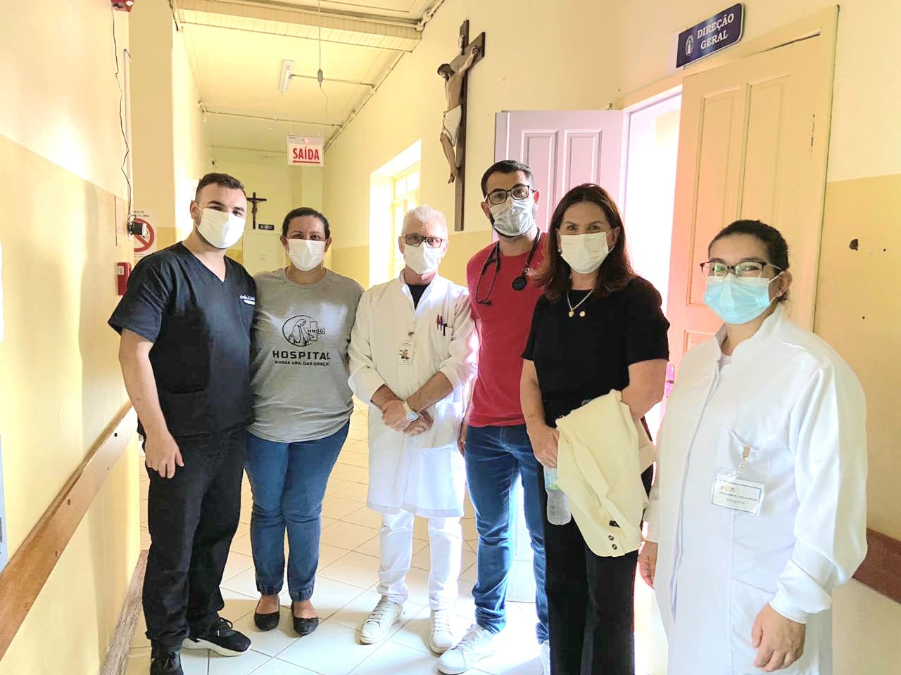 Seis profissionais da saúde e uma visitante posam para foto em corredor de hospital. Todos usam máscaras de proteção facial. Dois deles vestem jalecos brancos, um está com roupa preta, outro com roupa vermelha e estetoscópio no pescoço. Ao fundo, há uma porta aberta com a placa "Direção Geral" e uma placa de "Saída".