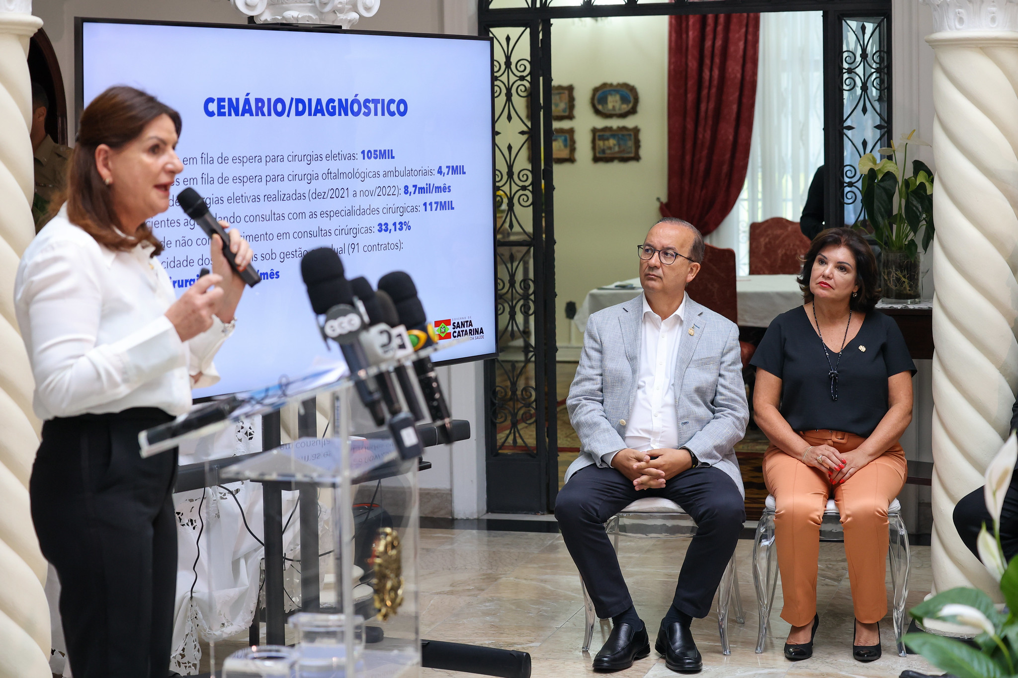 Mulher fala ao microfone em uma coletiva, ao lado de um telão que exibe dados do cenário e diagnóstico das filas de espera por cirurgias eletivas em Santa Catarina. À frente, estão microfones de emissoras de TV e, ao fundo, um homem e uma mulher estão sentados, acompanhando a apresentação.