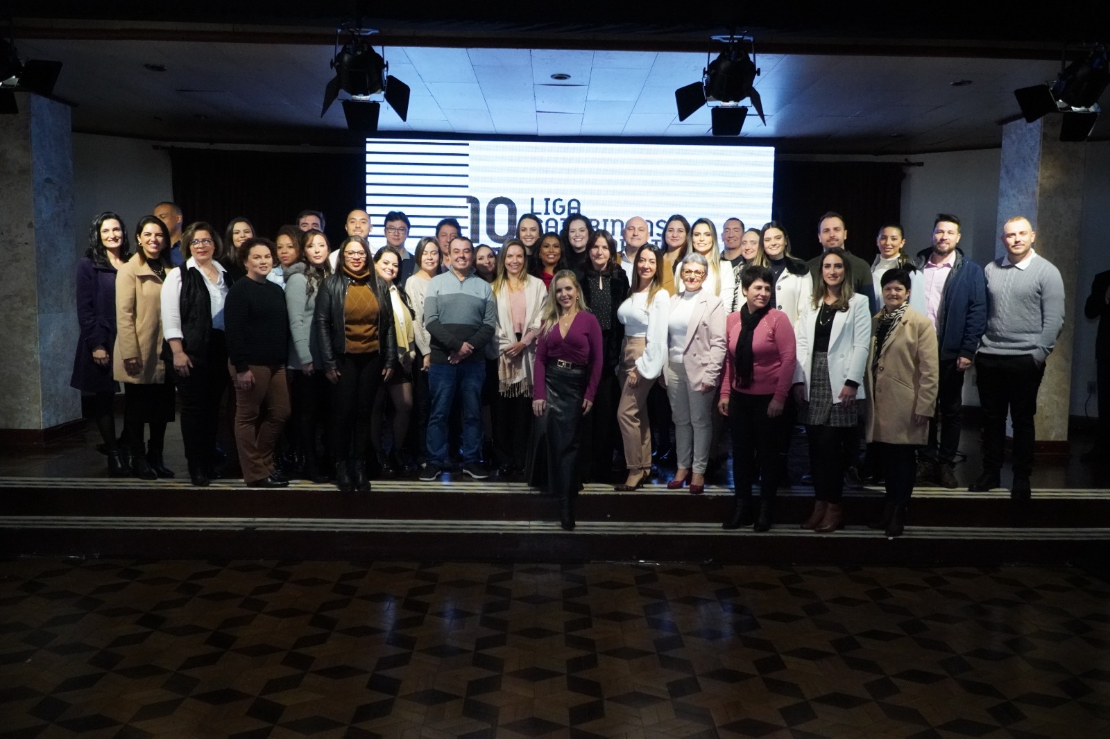 Grupo de profissionais posa em palco para foto comemorativa dos 10 anos da radioterapia no Hospital Tereza Ramos, com telão ao fundo exibindo o logotipo do evento.