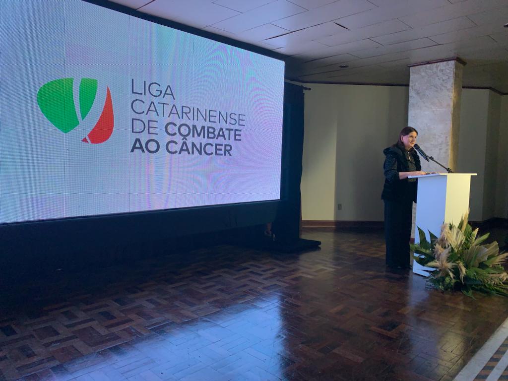 Mulher discursa em púlpito durante evento da Liga Catarinense de Combate ao Câncer, com grande telão exibindo o logotipo da organização ao fundo.