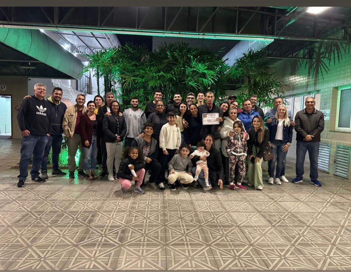 Grupo grande de pessoas, incluindo adultos, jovens e crianças, posa reunido em ambiente interno com piso de ladrilhos geométricos e plantas verdes ao fundo, iluminadas por luzes verdes. Algumas pessoas estão agachadas na frente, enquanto outras permanecem em pé atrás. Ao centro, uma pessoa segura um certificado. O ambiente é espaçoso, com teto metálico e tubulações aparentes. A atmosfera é informal e acolhedora, sugerindo um momento de confraternização ou homenagem.
