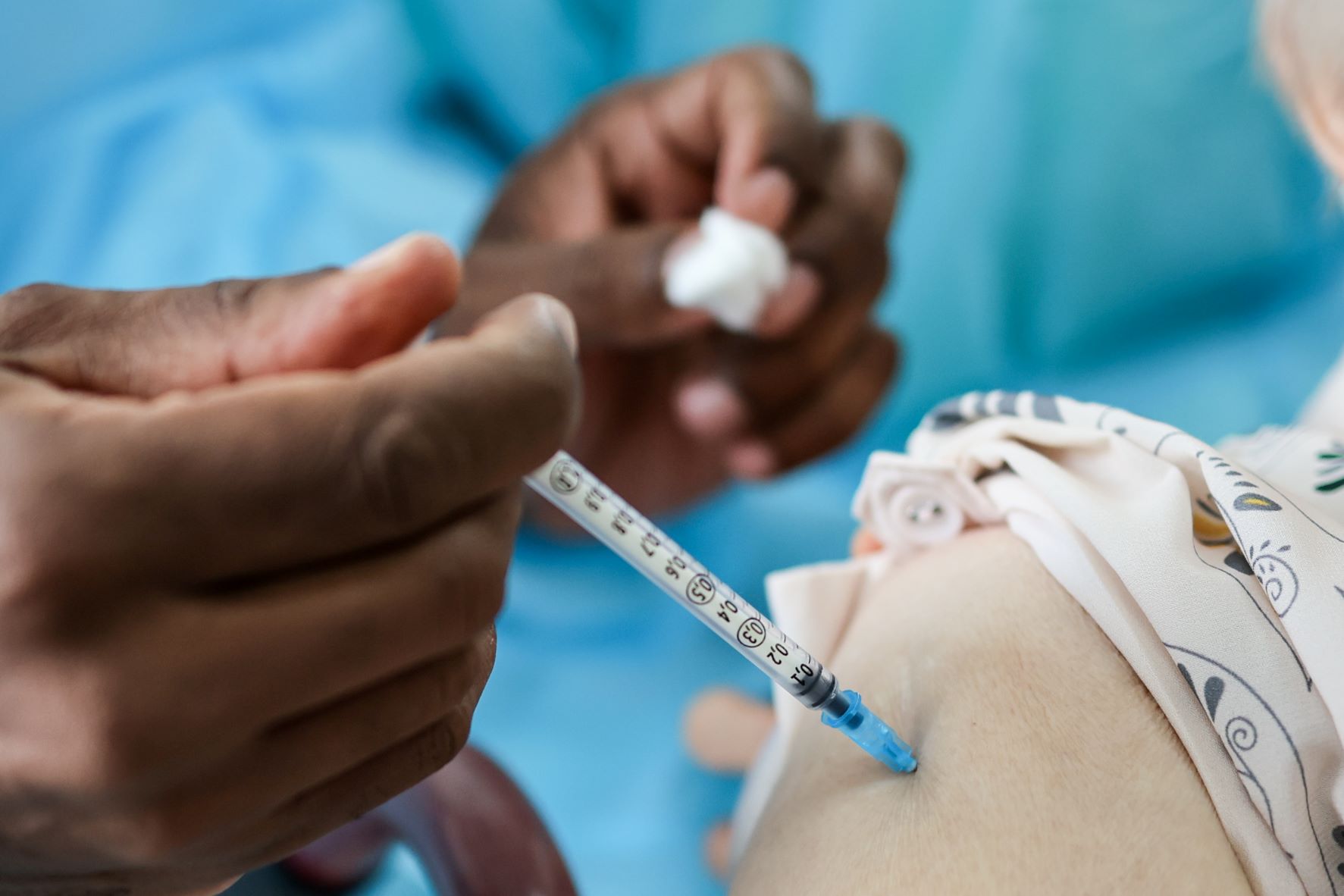 Close de uma aplicação de vacina no braço de uma pessoa. A seringa está inserida na pele e é segurada por um profissional de saúde com luvas. A outra mão segura um pedaço de algodão, provavelmente para uso após a injeção. A pessoa vacinada veste uma blusa clara com estampas coloridas. O fundo está desfocado, com tons de azul, indicando o uso de avental hospitalar.