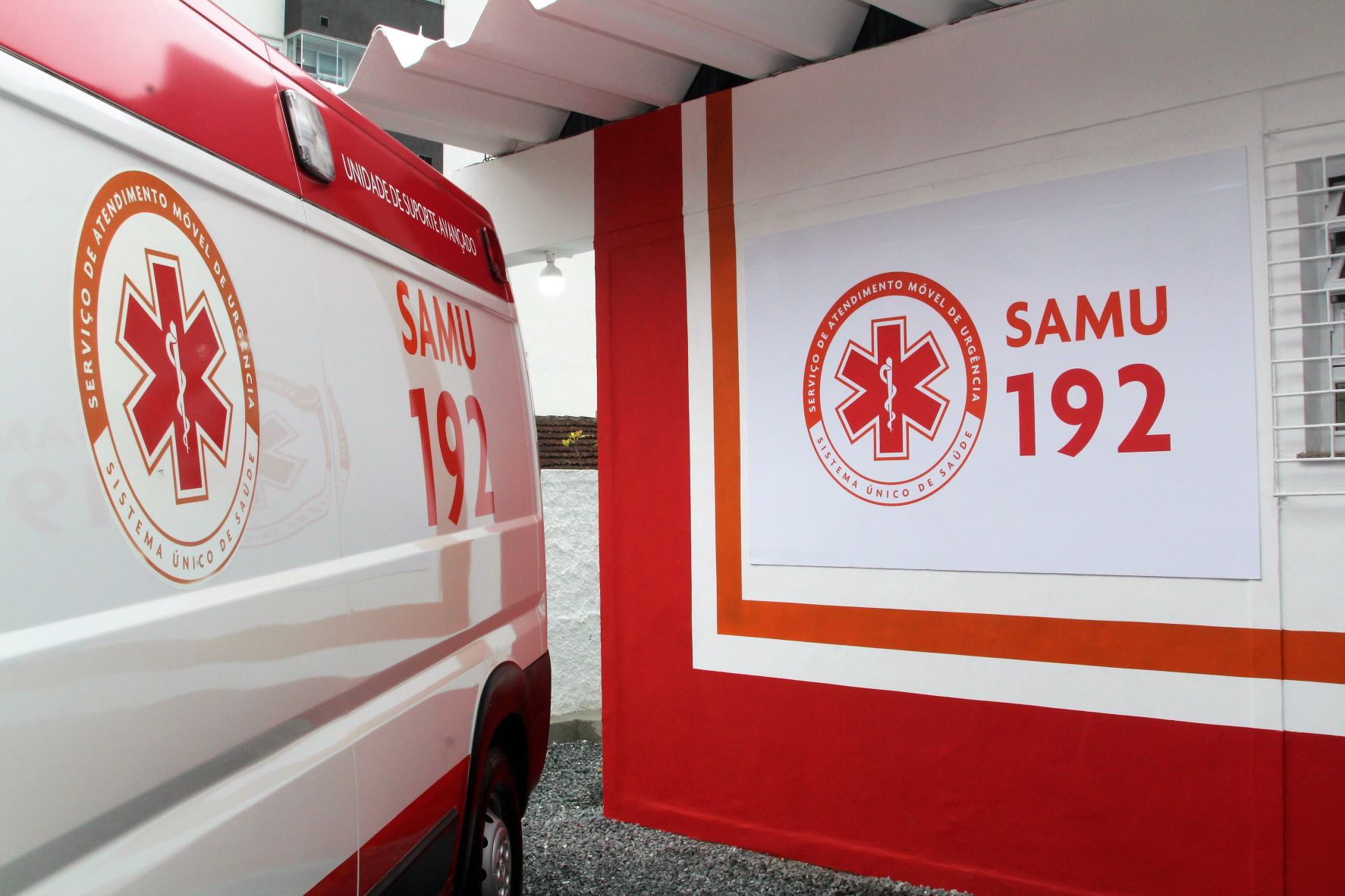 Imagem mostra uma ambulância do SAMU com a inscrição 'SAMU 192' e o logotipo do Serviço de Atendimento Móvel de Urgência, estacionada ao lado de uma parede com a mesma identidade visual. A parede é decorada com faixas nas cores branca, laranja e vermelha, e exibe o símbolo e o número de emergência do SAMU em destaque. A cena transmite a inauguração ou apresentação de uma nova unidade de atendimento.