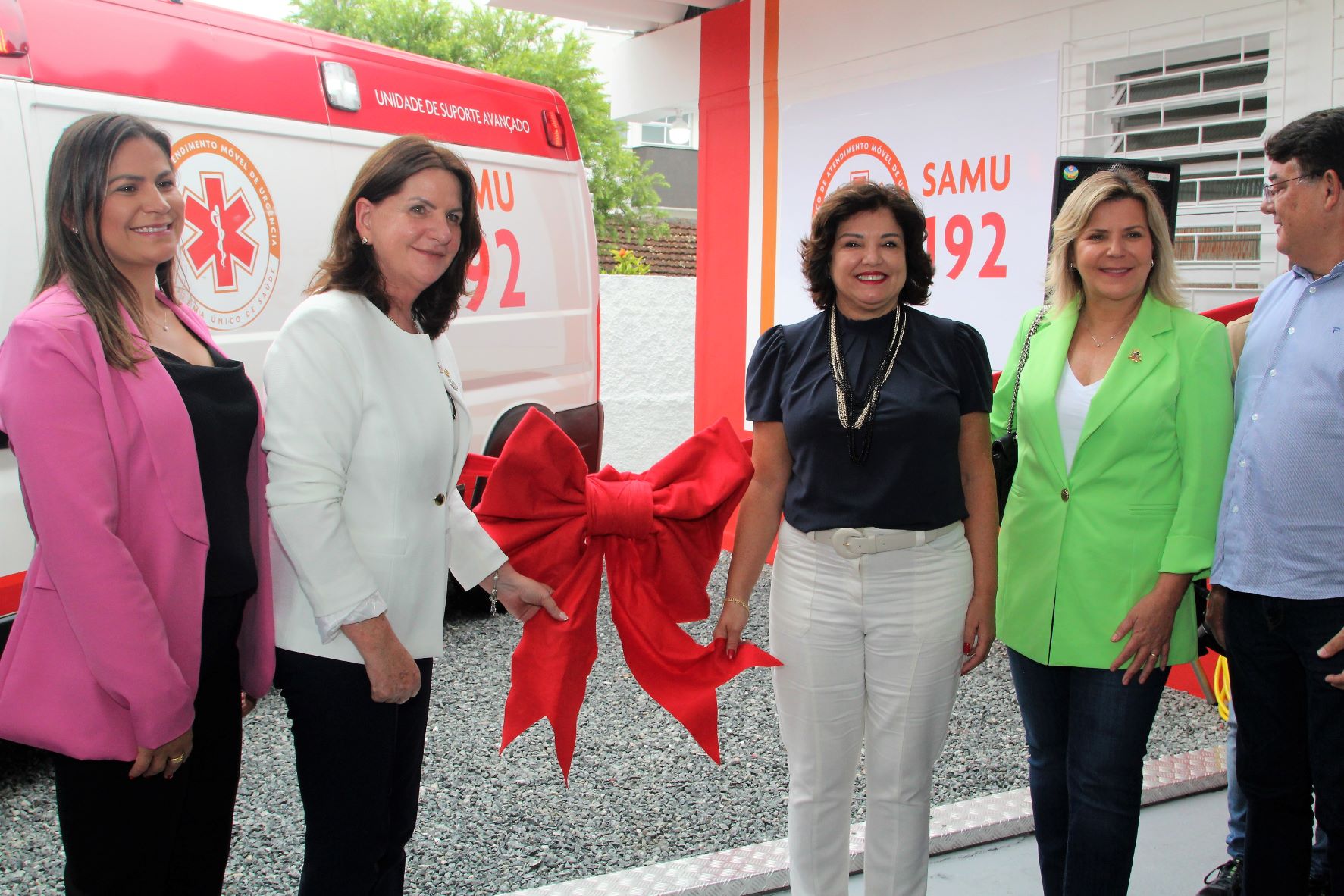 Cinco pessoas estão em pé em frente a uma ambulância do SAMU com o número 192 visível. Duas mulheres ao centro seguram um grande laço vermelho simbólico, sugerindo a inauguração de um novo serviço ou equipamento de saúde. À esquerda, uma mulher veste blazer rosa; ao centro, outra de blazer branco; ao lado direito, uma mulher com blusa escura e calça branca, outra com blazer verde, e um homem de camisa azul claro. Todos estão sorrindo. Ao fundo, o letreiro indica "Unidade de Suporte Avançado – SAMU".