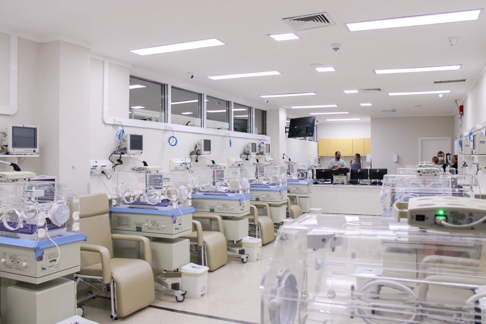 Ambiente moderno de UTI Neonatal com diversas incubadoras alinhadas lado a lado, cada uma equipada com monitores e aparelhos médicos. O espaço é bem iluminado, com paredes brancas e cadeiras de apoio ao lado de cada incubadora. Profissionais da saúde conversam ao fundo, próximos à central de enfermagem.