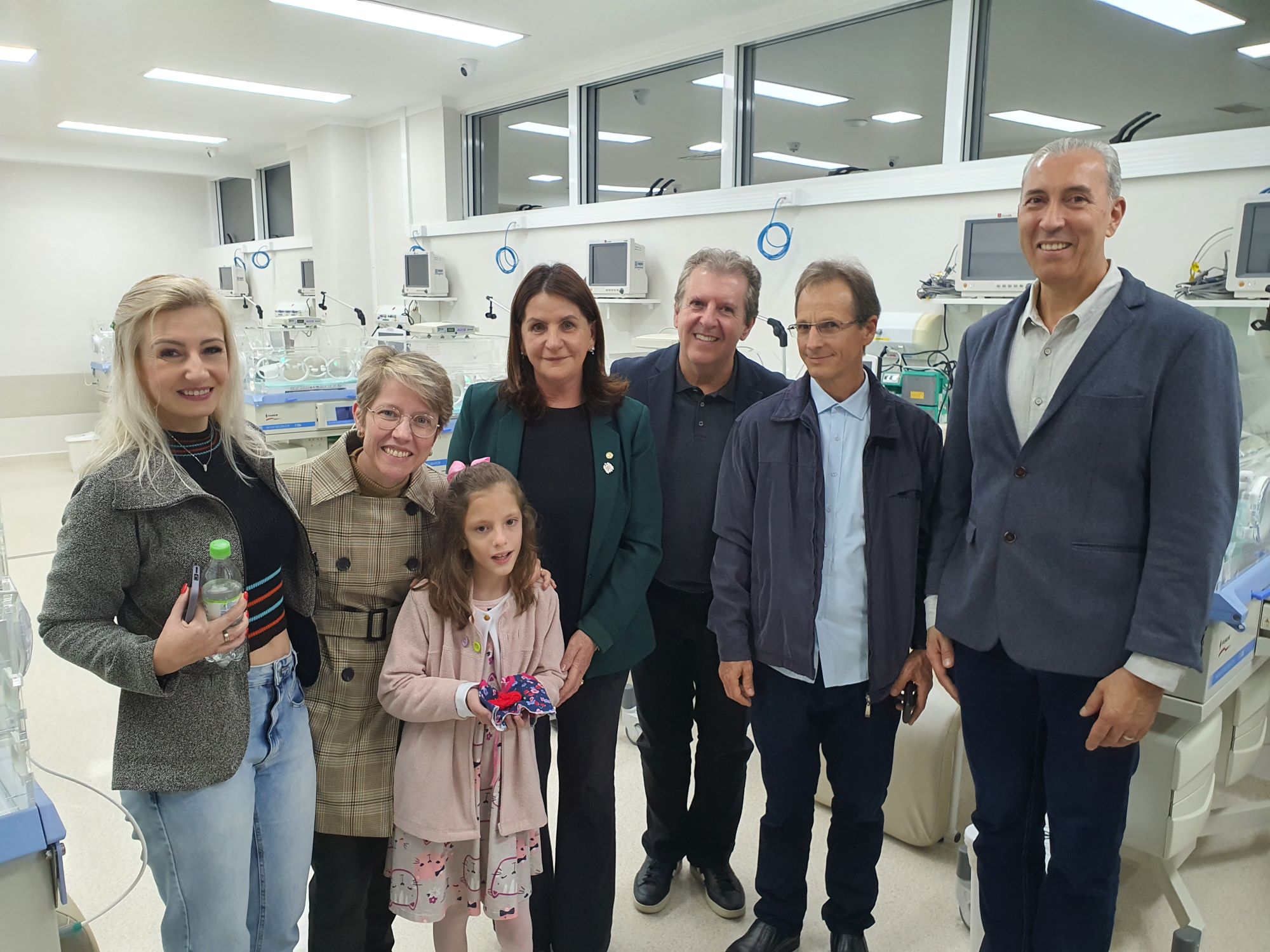 Grupo de sete pessoas posando sorridente em uma unidade de terapia intensiva neonatal (UTI neonatal), com incubadoras e equipamentos hospitalares ao fundo. Entre elas, uma criança em destaque ao centro, cercada por adultos. O ambiente é moderno, bem iluminado e limpo, com equipamentos médicos organizados ao longo da parede.