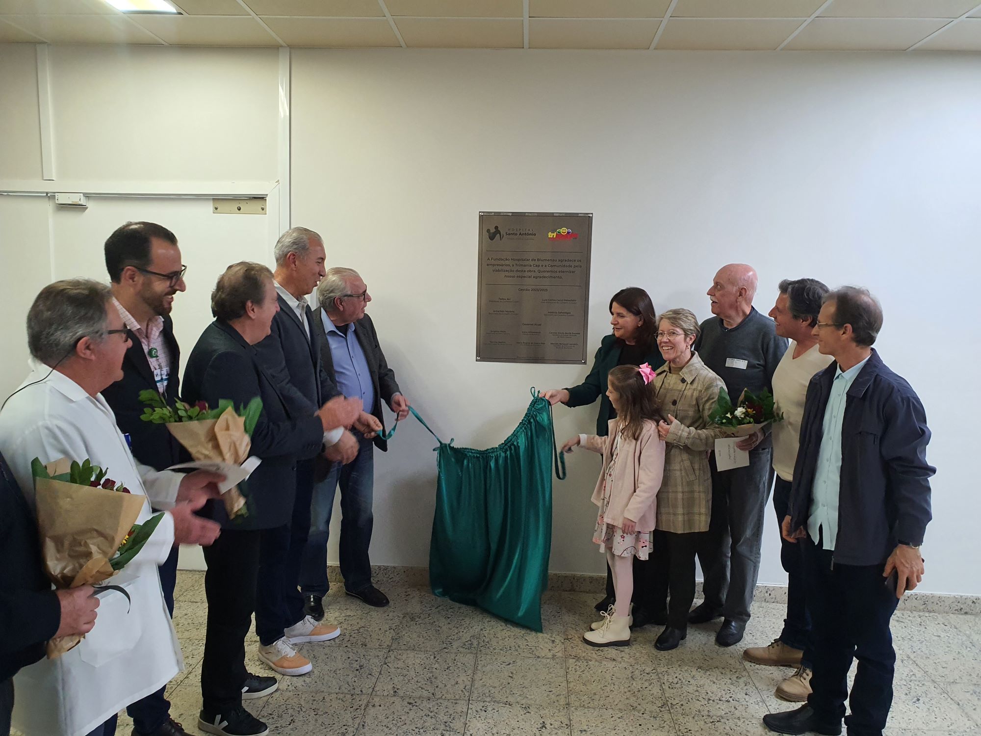 Grupo de pessoas reunidas em frente a uma placa comemorativa durante uma cerimônia de inauguração. Ao centro, uma mulher e uma criança seguram um tecido verde que foi usado para revelar a placa. Algumas pessoas seguram buquês de flores, e todos estão sorrindo em um ambiente interno com piso claro e teto de gesso.