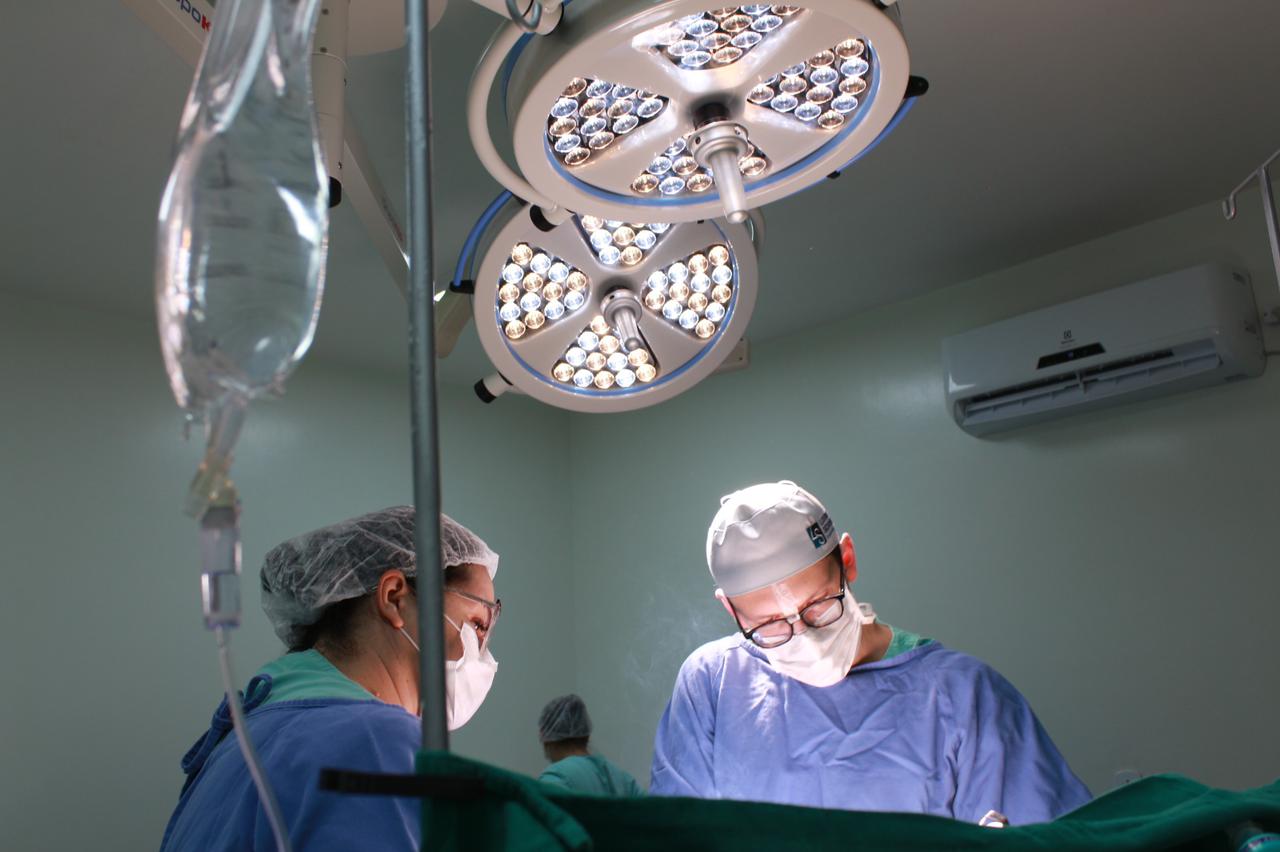 Equipe médica realizando um procedimento cirúrgico em centro cirúrgico moderno, iluminado por refletores cirúrgicos LED. Os profissionais utilizam roupas e máscaras esterilizadas, com foco concentrado no procedimento.