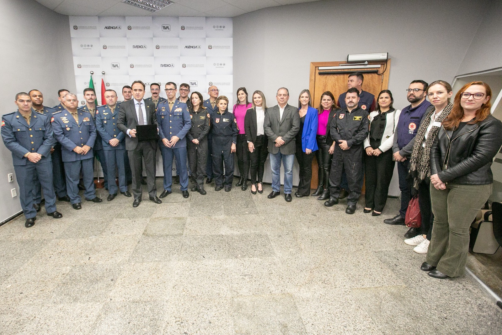Grupo de profissionais da segurança pública e da saúde, incluindo bombeiros militares, socorristas do SAMU, policiais e autoridades civis, reunidos em uma cerimônia ou encontro institucional em ambiente fechado com fundo institucional e bandeiras.