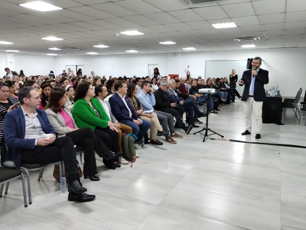 Auditório cheio com profissionais atentos a uma apresentação. Um palestrante, de terno escuro e calça clara, fala ao microfone em pé próximo a um projetor. Na primeira fila, diversas autoridades e representantes acompanham a palestra.