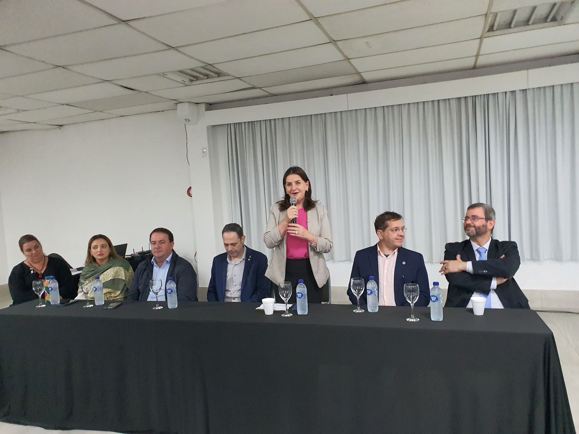 Autoridades e profissionais da saúde sentados à mesa durante evento institucional em auditório. Uma mulher ao centro, em pé com microfone, faz um discurso enquanto os demais a observam. Garrafas de água e taças estão sobre a mesa coberta com toalha preta.
