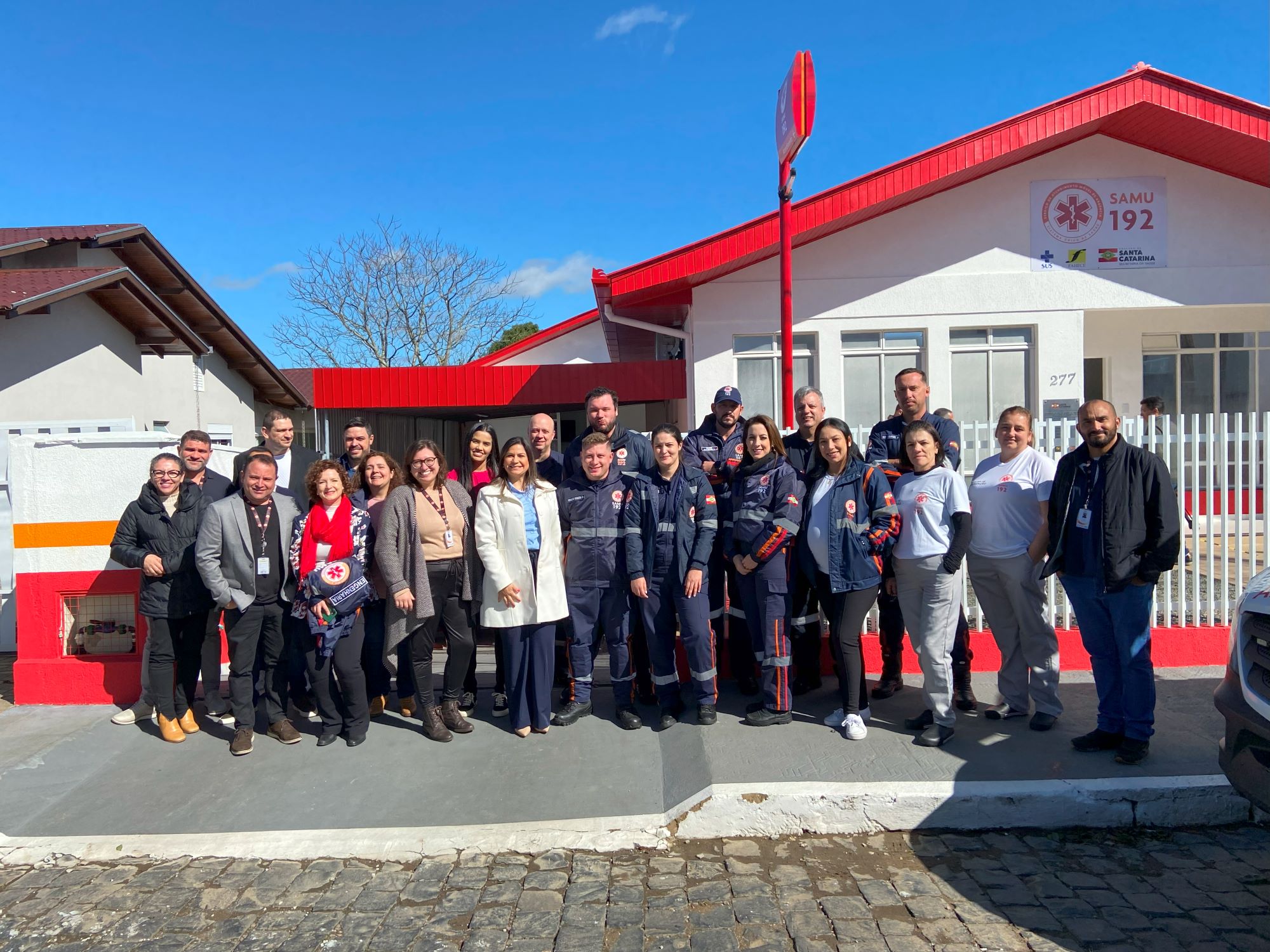 Grupo de profissionais do SAMU e representantes da saúde posando em frente à base do SAMU, com prédio branco e vermelho ao fundo. Todos sorridentes, em dia ensolarado, celebrando ou registrando visita técnica.