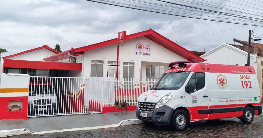 Base do SAMU com fachada branca e detalhes vermelhos, em frente à qual estão estacionadas duas ambulâncias do serviço de atendimento móvel de urgência, uma dentro do pátio e outra na rua. Céu nublado e um cachorro na calçada.