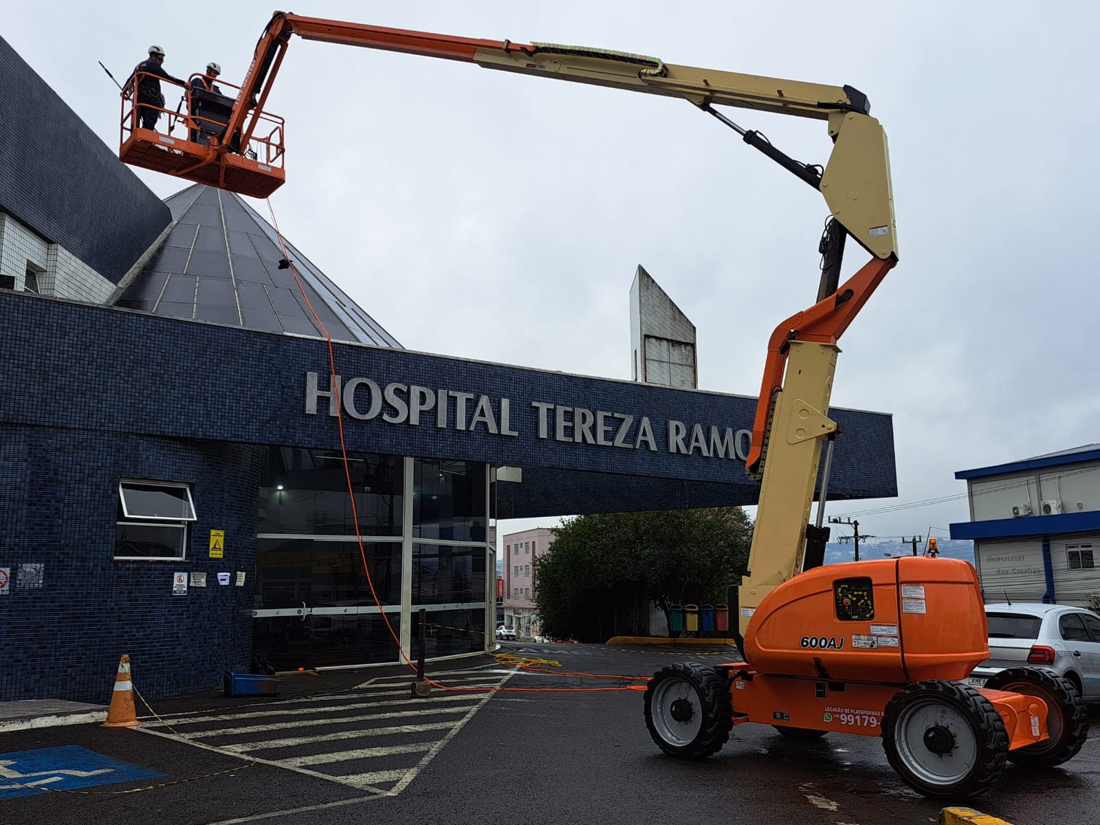 Dois trabalhadores em plataforma elevatória realizam manutenção na estrutura envidraçada do Hospital Tereza Ramos, em dia nublado. Equipamento hidráulico de cor laranja está posicionado na entrada do hospital.