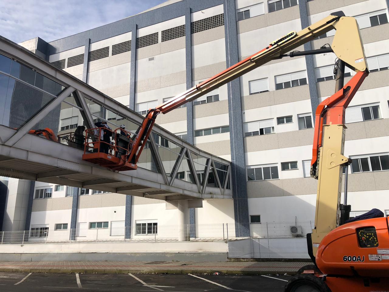 Trabalhadores utilizam plataforma elevatória laranja para realizar manutenção em passarela envidraçada suspensa, conectada a edifício hospitalar de vários andares, em dia claro.