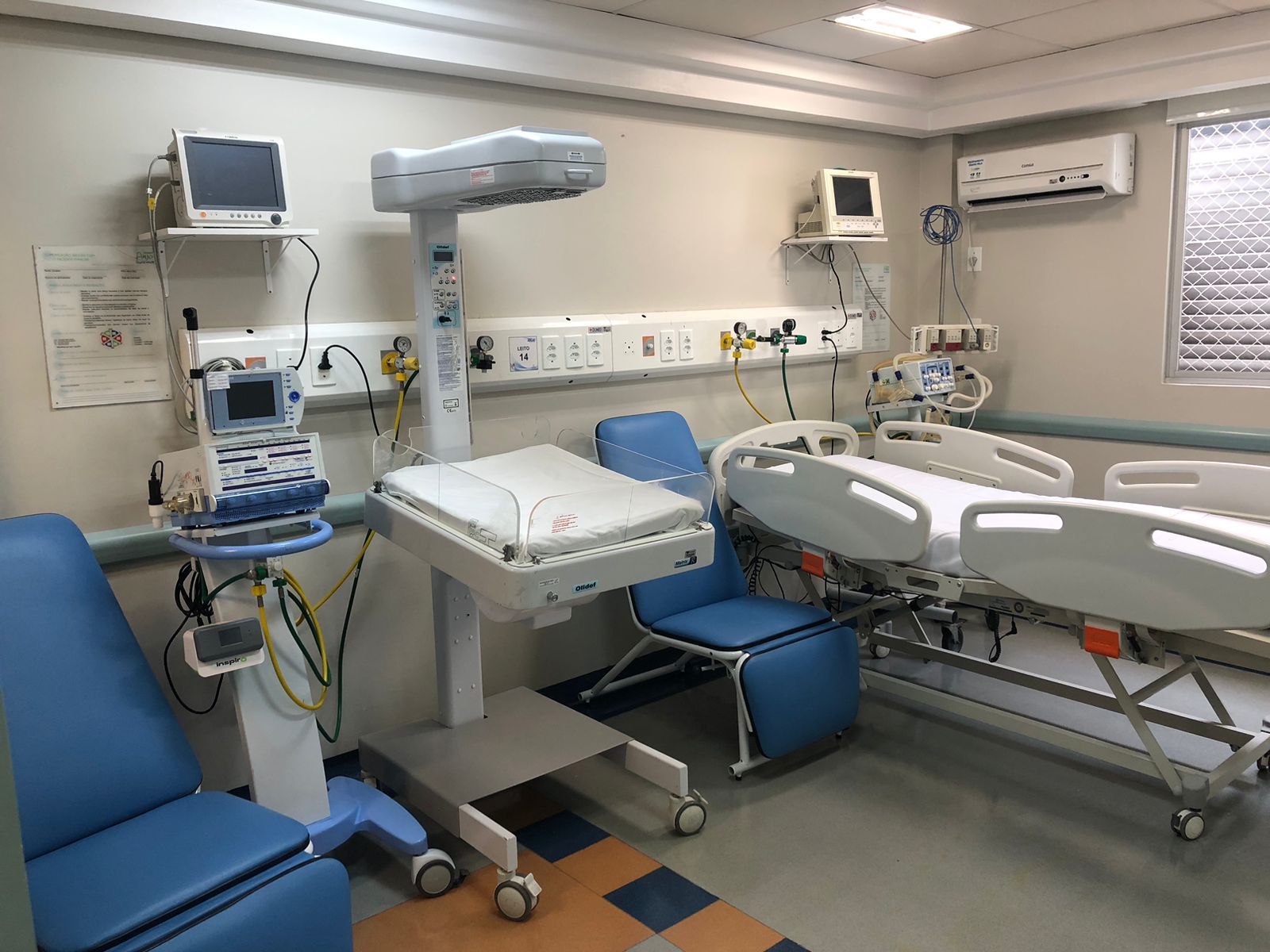 Quarto hospitalar equipado com cama moderna, incubadora neonatal, poltronas reclináveis e diversos monitores e aparelhos médicos fixados à parede. Ambiente limpo e bem iluminado, com infraestrutura de UTI.