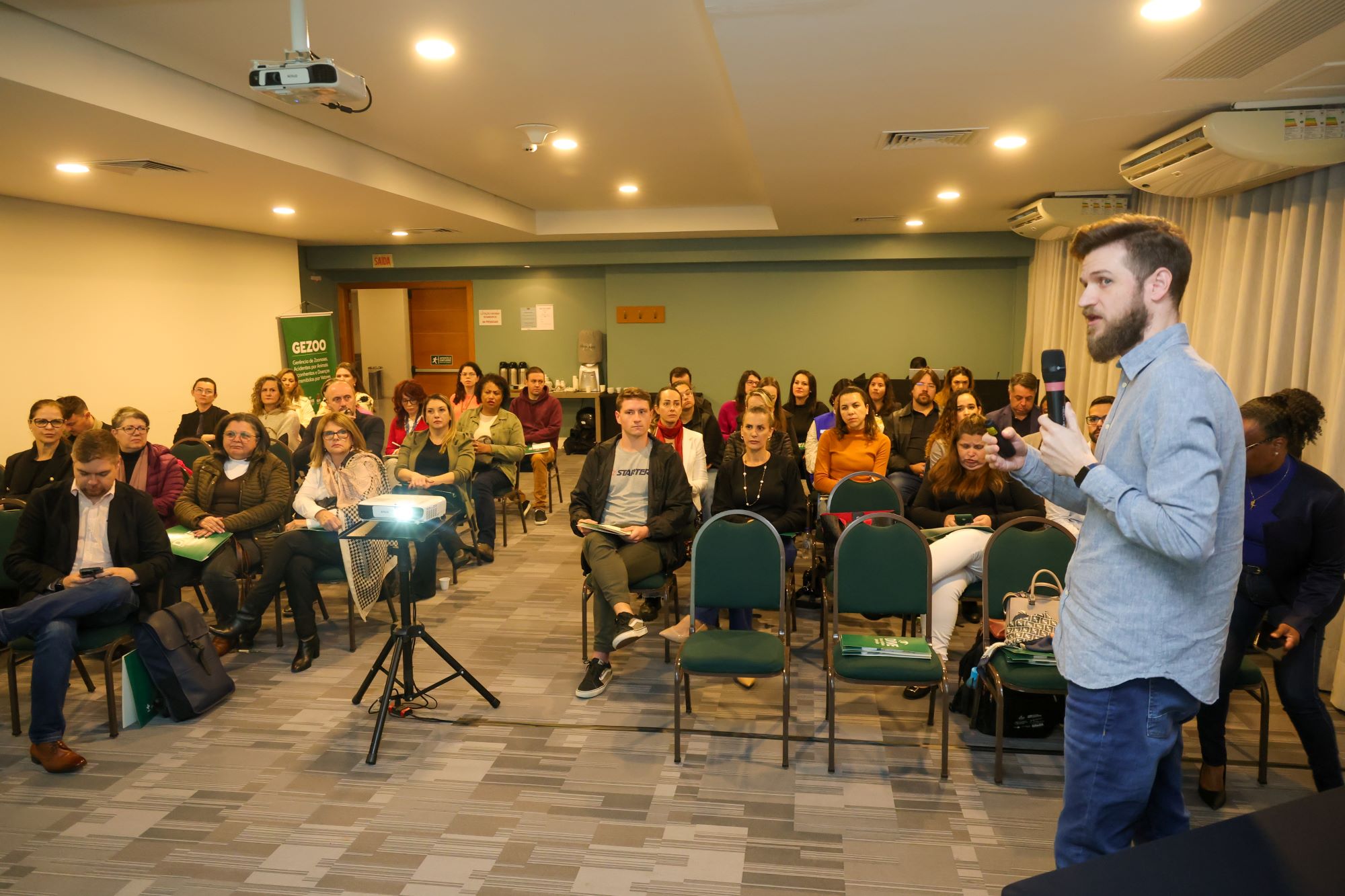Homem segurando microfone e palestrando para um auditório cheio de pessoas sentadas, com projetor ligado e banner do GEZOO ao fundo.