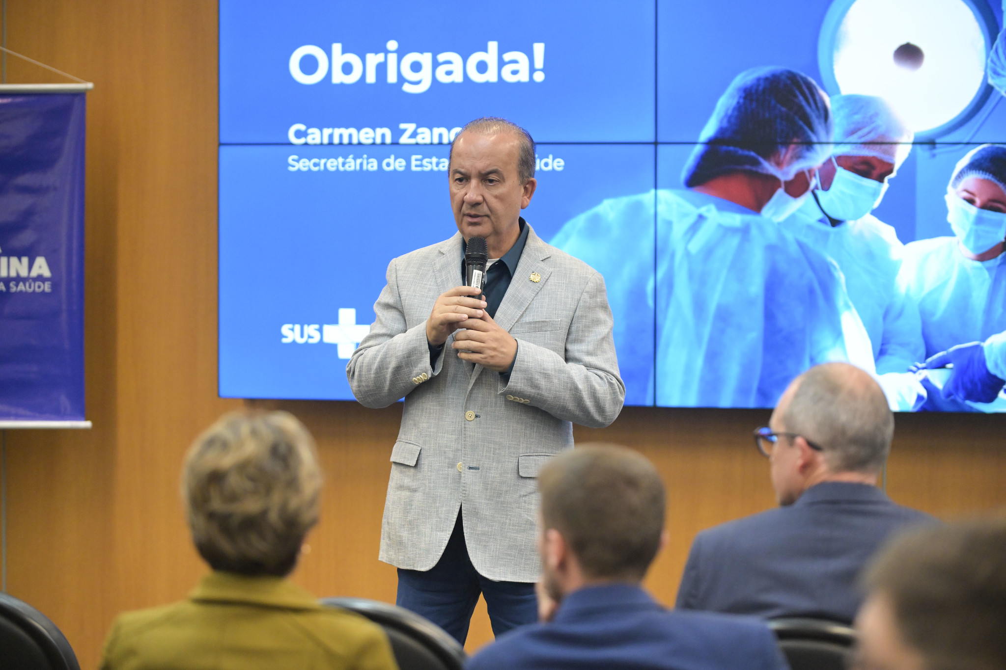 Profissional de saúde em trajes cirúrgicos azuis utiliza um equipamento chamado VeinViewer para localizar veias em um recém-nascido que está deitado em uma incubadora neonatal. O aparelho projeta uma luz verde fluorescente sobre a pele do bebê, revelando a localização das veias. A profissional está usando luvas e tocando cuidadosamente o braço do bebê, que está parcialmente coberto com fralda.