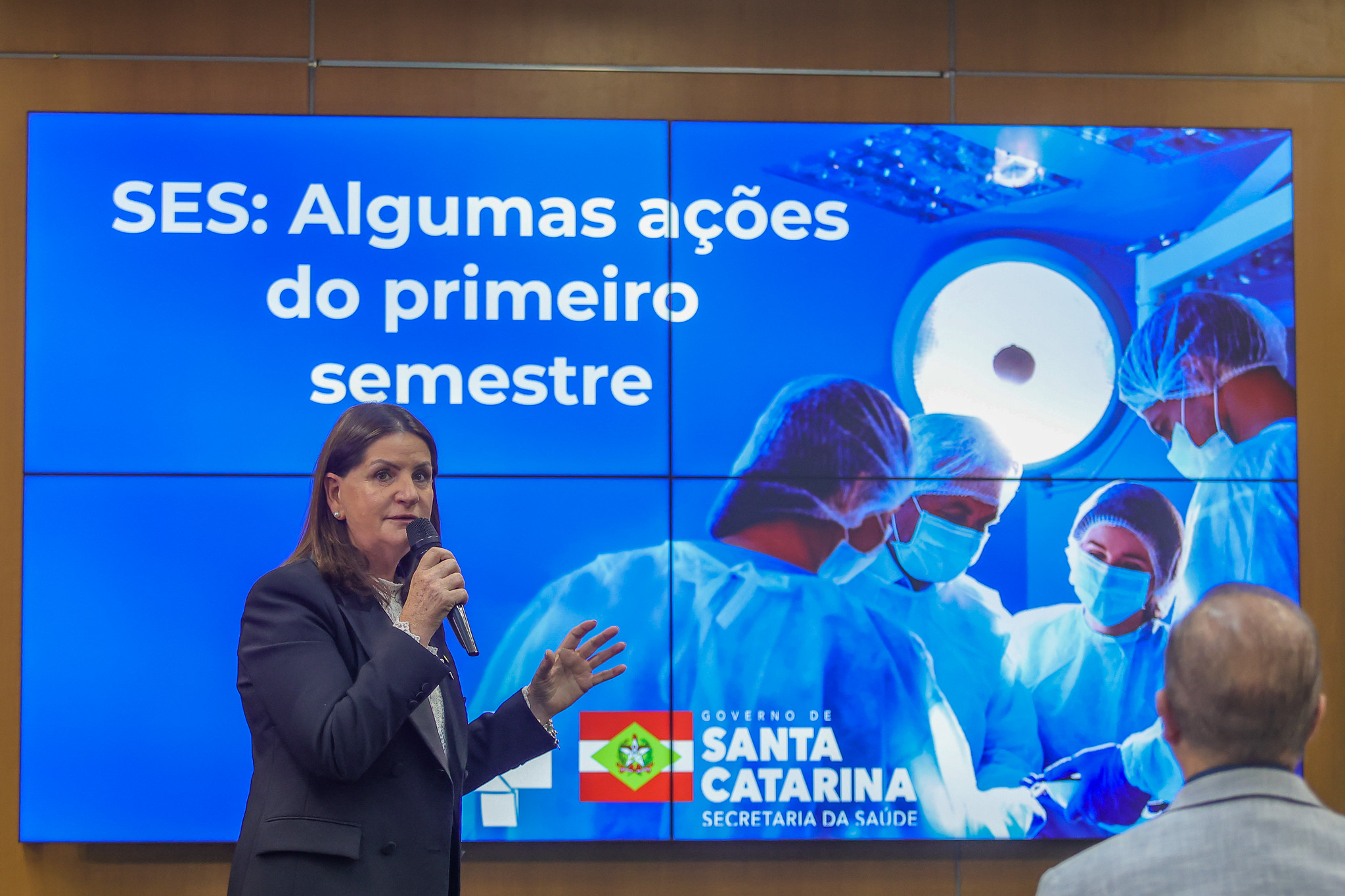 Profissional da saúde vestindo uniforme cirúrgico azul, touca branca, máscara rosa e óculos, segura um endoscópio apontado para a câmera. A pessoa está de frente, com expressão séria, demonstrando preparo para o manuseio do equipamento médico, utilizado em procedimentos de diagnóstico e tratamento por imagem interna. O fundo é uma parede clara, sem distrações visuais.