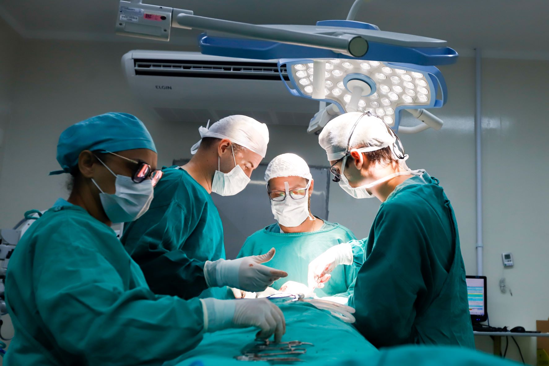 Equipe médica composta por quatro profissionais realiza uma cirurgia em um centro cirúrgico. Todos estão vestidos com roupas estéreis verdes, máscaras, luvas e toucas cirúrgicas. Eles trabalham em conjunto, focados no procedimento, sob uma luz cirúrgica intensa. Instrumentos cirúrgicos estão organizados sobre o paciente, coberto por campos cirúrgicos. Ao fundo, vê-se parte de um monitor e aparelhos hospitalares, indicando ambiente altamente controlado e técnico.