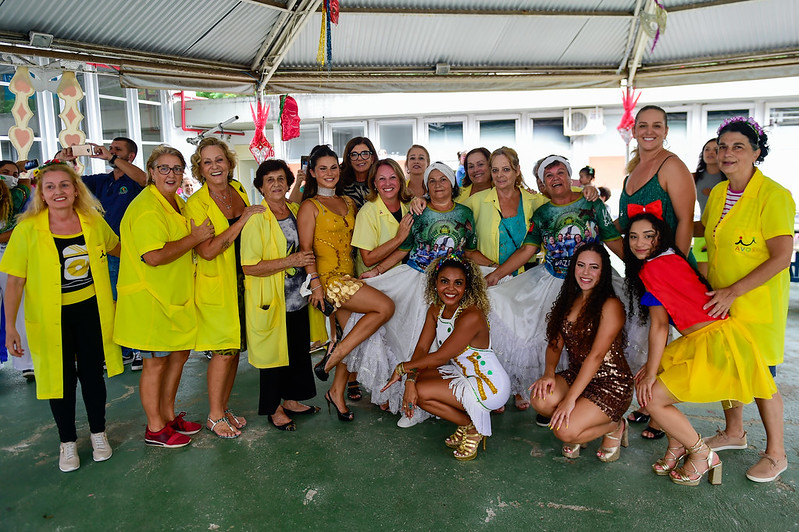 Grupo de mulheres posa sorridente durante uma confraternização com tema carnavalesco em ambiente coberto. Algumas estão vestidas com fantasias coloridas, outras com jalecos amarelos do projeto 'A Voz do Coração'. O clima é de alegria e celebração, com decoração festiva ao fundo.
