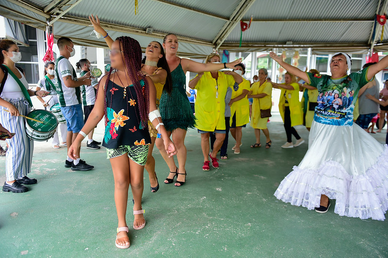 Mulheres e uma menina participam de uma animada roda de samba em um espaço coberto e decorado para o carnaval. À esquerda, músicos com instrumentos de percussão acompanham a dança. Algumas participantes usam fantasias coloridas, outras vestem jalecos amarelos do projeto 'A Voz do Coração'. O ambiente é de celebração e inclusão.