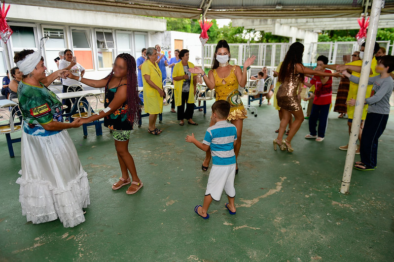 Crianças, adultos e idosos dançam juntos em uma área coberta decorada com enfeites de carnaval. À esquerda, uma mulher com roupa branca típica do samba segura as mãos de uma menina sorridente. Ao centro, uma passista fantasiada interage com um menino pequeno. Ao fundo, voluntárias com jalecos amarelos e outras pessoas participam da confraternização em clima de alegria e inclusão.