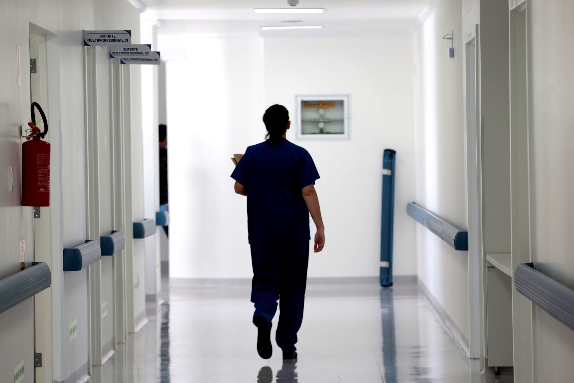 Profissional de saúde de costas, usando uniforme azul, caminha por um corredor hospitalar iluminado e limpo. À esquerda, há portas com placas indicando "Suporte Multiprofissional" e um extintor de incêndio preso à parede.