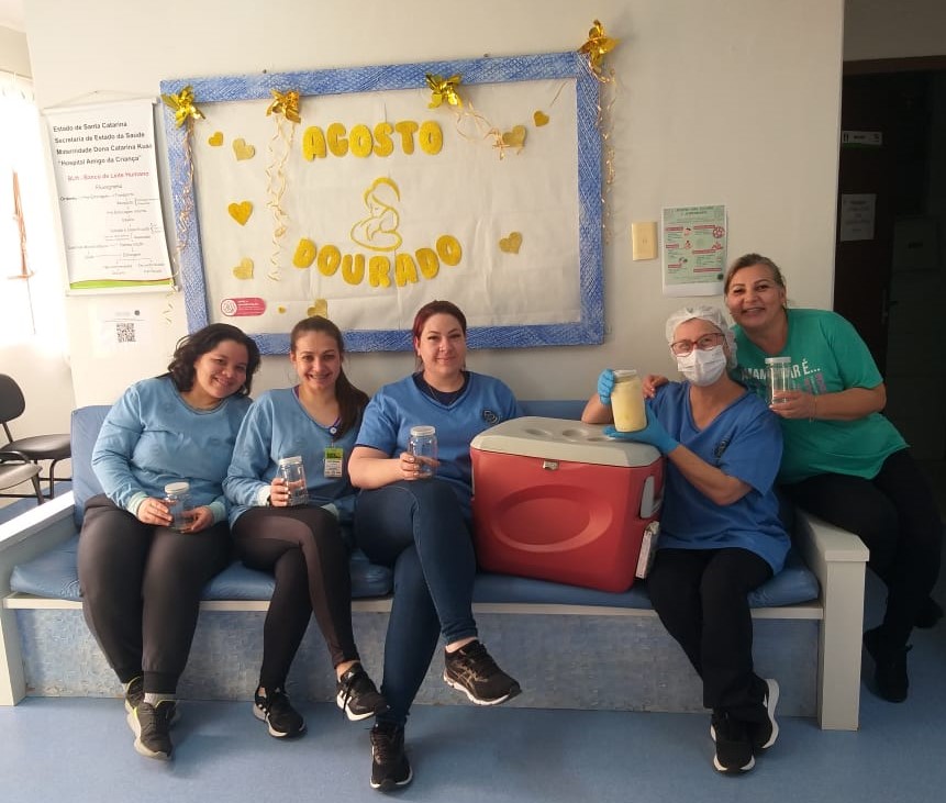 Cinco mulheres sentadas em um banco hospitalar, sorrindo e segurando potes de vidro com leite humano. Ao centro, há uma caixa térmica vermelha com tampa cinza. Atrás delas, um painel decorativo com o tema "Agosto Dourado", que destaca a amamentação, adornado com estrelas douradas e desenhos de corações. Uma das profissionais usa touca, máscara e luvas, segurando um frasco de leite.