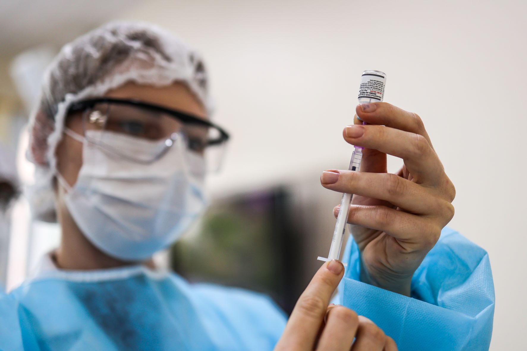 Profissional de saúde com máscara, touca, óculos de proteção e avental azul prepara uma seringa com uma dose de vacina, segurando o frasco com cuidado. A imagem está em foco na seringa e na ampola, enquanto o rosto da profissional aparece desfocado ao fundo.