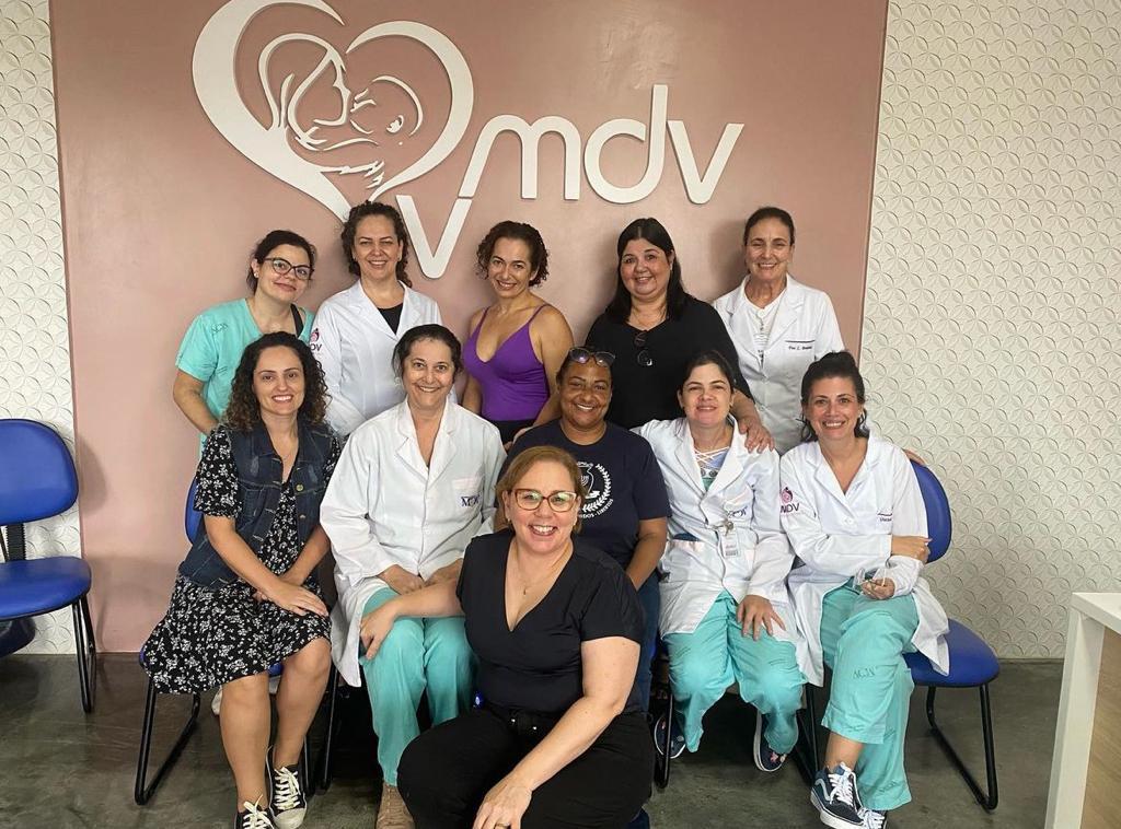 Grupo de profissionais de saúde, incluindo enfermeiras, técnicas e colaboradoras, posando sorridentes em frente ao logotipo do Banco de Leite Humano MDV, demonstrando união e dedicação ao cuidado materno-infantil.