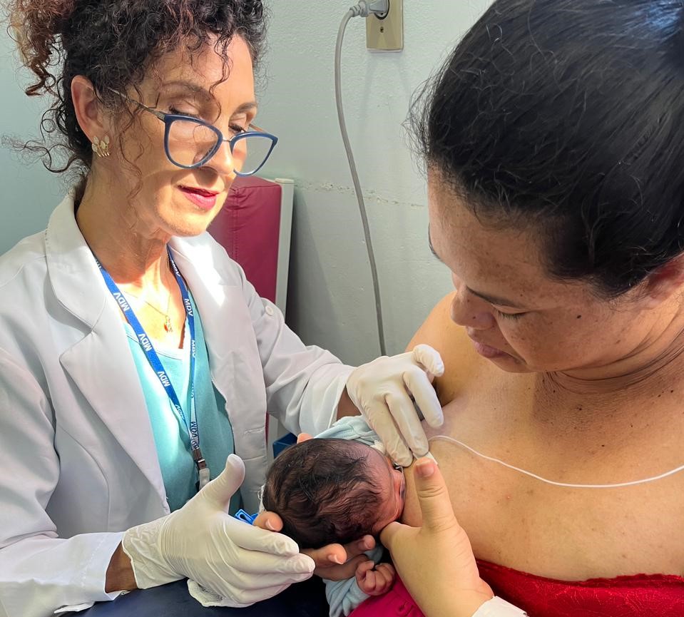 Profissional de saúde com jaleco branco, óculos e luvas orienta mãe durante o uso da técnica sonda-dedo para amamentação de recém-nascido. A mãe segura o bebê junto ao peito, enquanto uma sonda fina é usada para alimentação complementar.