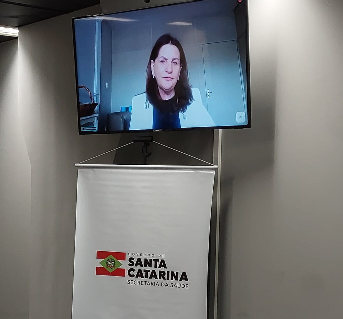 Tela exibindo mulher em videoconferência, com banner abaixo contendo o logotipo do Governo de Santa Catarina e a inscrição "Secretaria da Saúde". A mulher usa blazer branco e fone de ouvido.