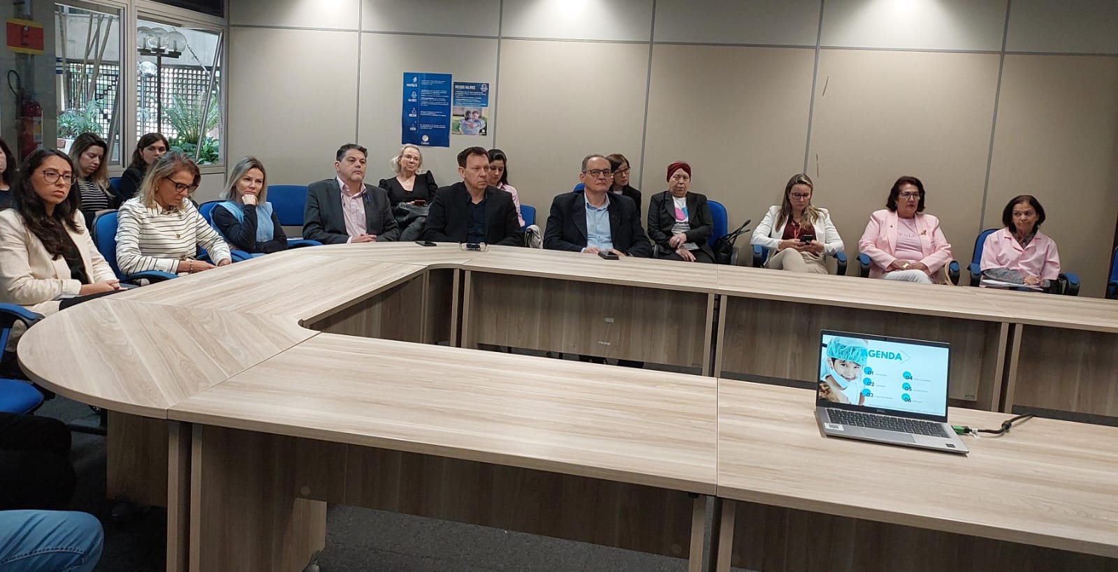 Grupo de pessoas sentadas ao redor de uma mesa em formato de U durante uma reunião. Na frente, há um notebook exibindo uma apresentação com o título "AGENDA". A maioria das mulheres veste roupas em tons de rosa, sugerindo uma campanha ou evento temático.