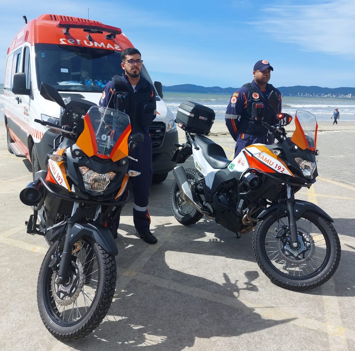 Fachada de um prédio hospitalar de vários andares, com janelas alinhadas e diversas unidades de ar-condicionado instaladas. A entrada principal tem uma cobertura verde, com uma área de acesso para veículos. Em frente ao prédio, há um jardim bem cuidado com árvores, arbustos, gramado e um caminho de pedra por onde circulam pessoas e um carro estacionado.