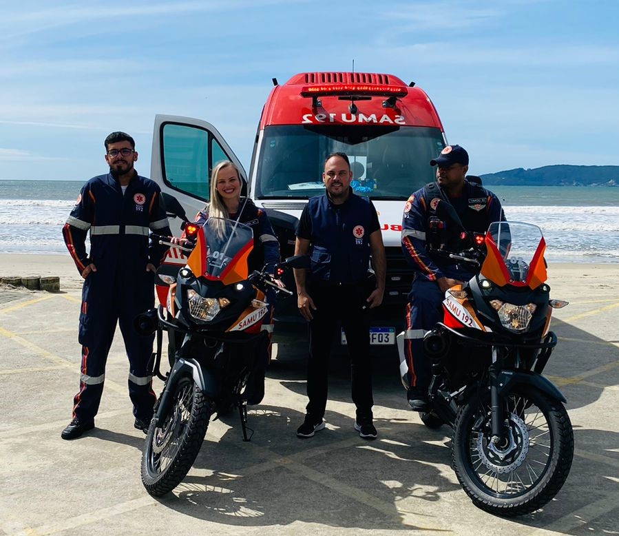 Quatro profissionais do SAMU posam em frente a uma ambulância estacionada à beira-mar, em um dia ensolarado. Dois deles estão sobre motolâncias com o logotipo SAMU 192, usando uniformes escuros com faixas reflexivas. A ambulância vermelha e branca está com a porta aberta e ao fundo vê-se o mar e montanhas. A imagem transmite prontidão e mobilização da equipe de emergência.