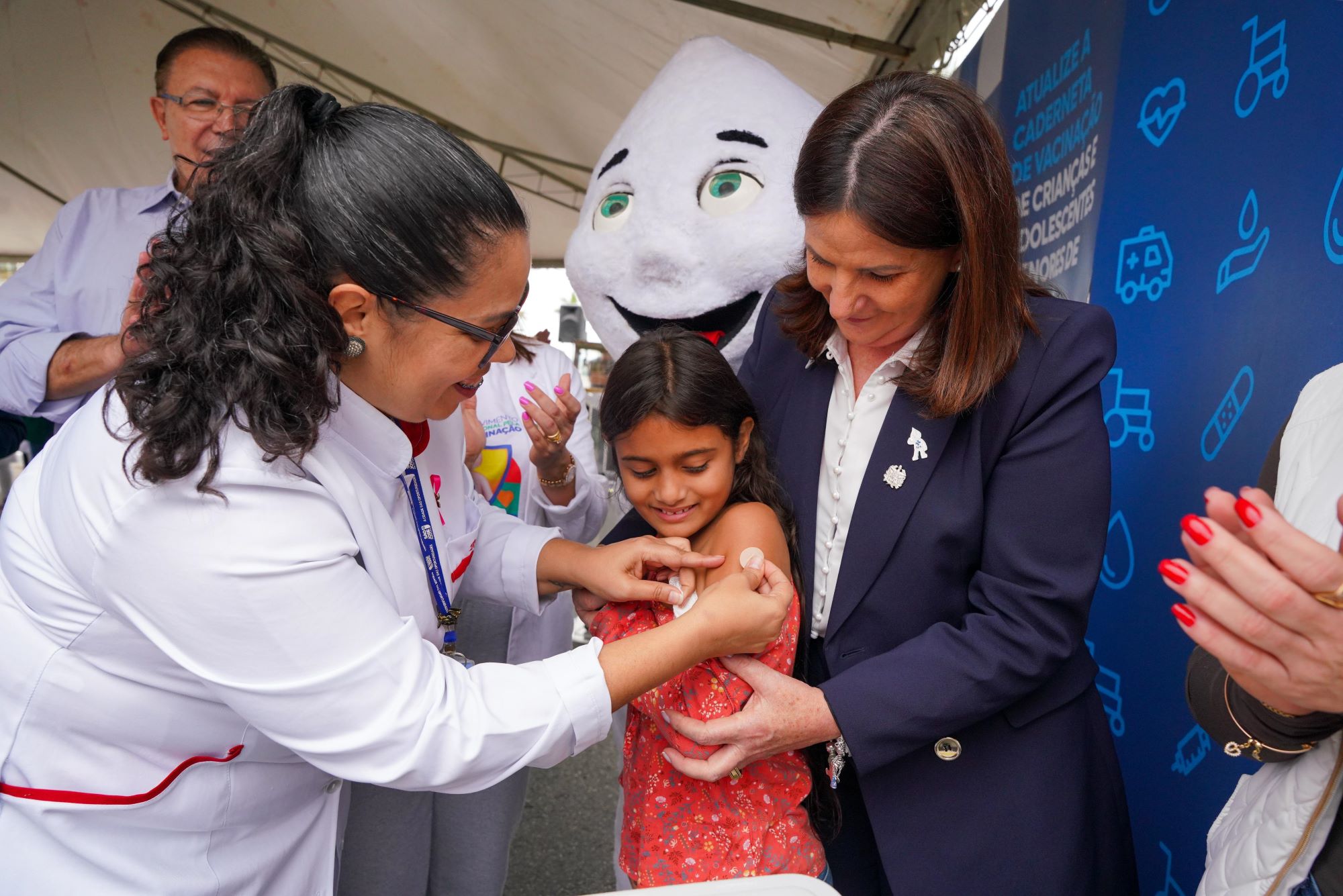 Profissional de saúde aplica vacina em uma criança sorridente, acompanhada por uma mulher que a segura carinhosamente. Ao fundo, está o personagem Zé Gotinha, símbolo das campanhas de vacinação no Brasil, em um ambiente festivo e educativo.
