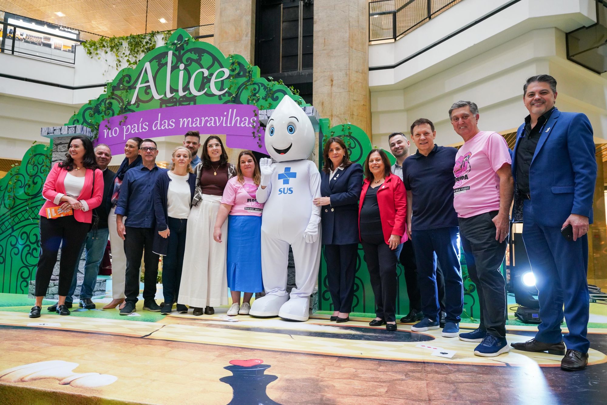 Grupo de pessoas posa ao lado do Zé Gotinha em frente a um painel com o tema "Alice no País das Maravilhas", em um ambiente decorado, promovendo campanha de vacinação.
