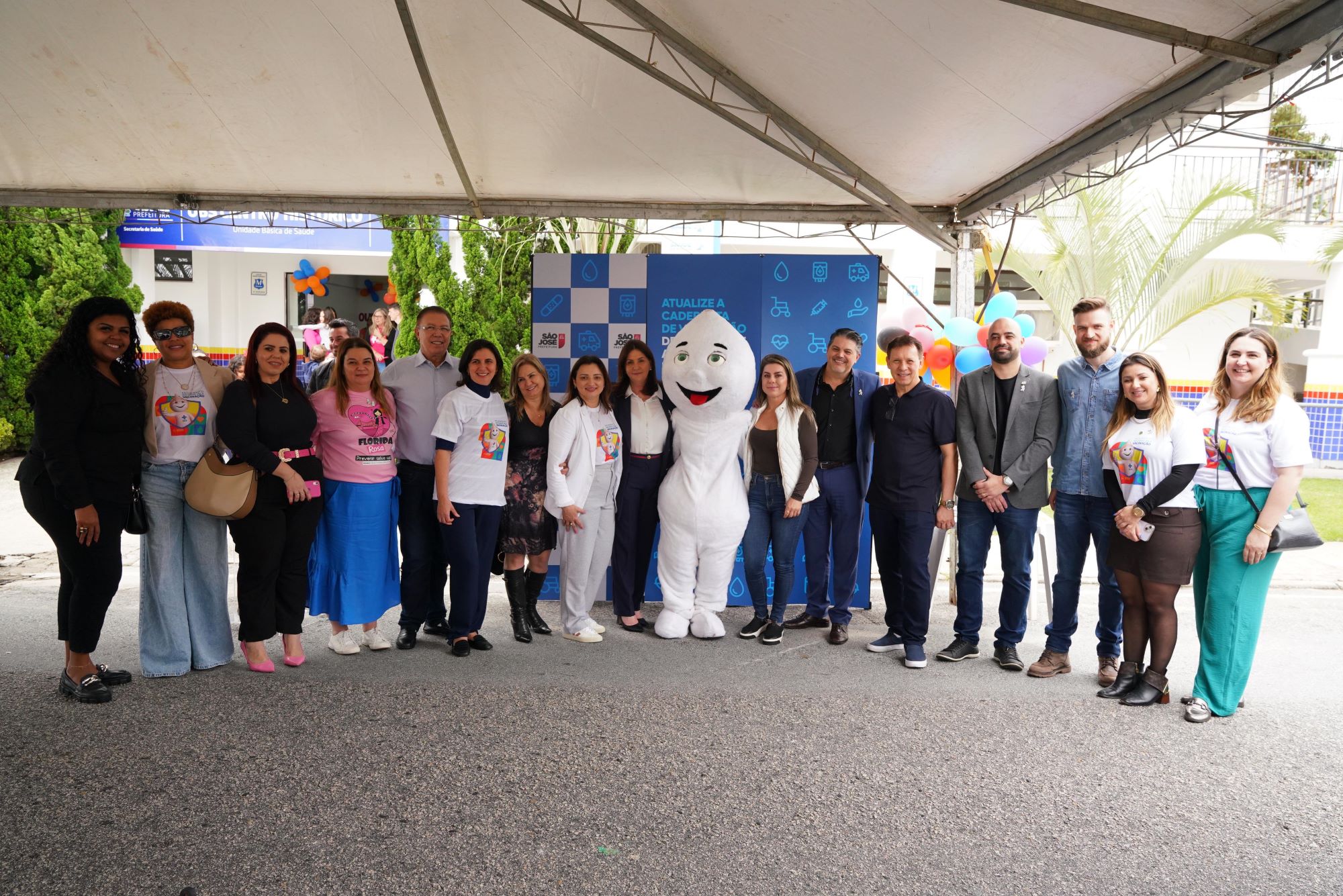 Grupo de profissionais e autoridades posam ao lado do Zé Gotinha em frente a unidade básica de saúde, sob uma tenda, com painel e balões ao fundo, promovendo a campanha de vacinação.