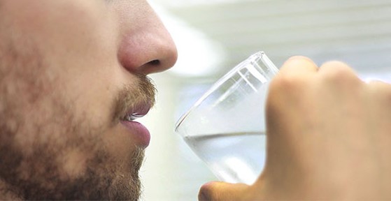 Homem com barba rala visto em close bebendo um copo de água transparente, com fundo desfocado.