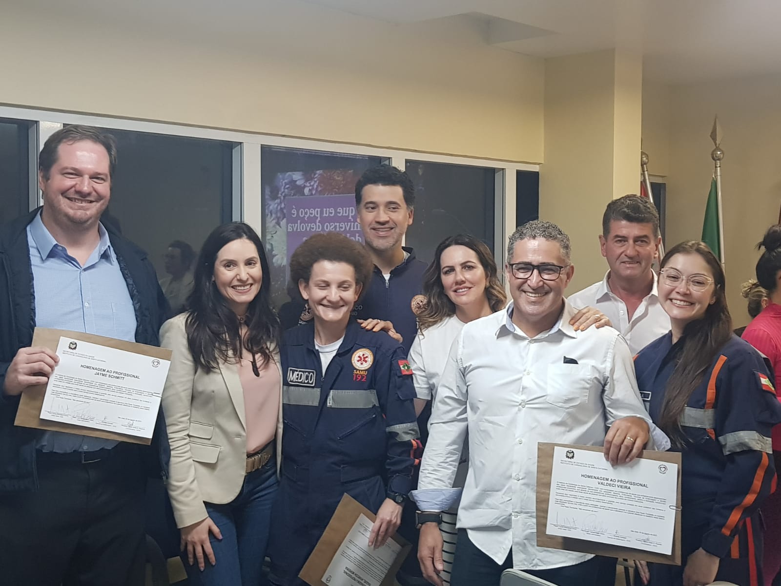Grupo de profissionais do SAMU e autoridades posam sorridentes para foto em ambiente interno. Três pessoas seguram certificados de homenagem, e todos demonstram alegria e orgulho. Ao fundo, bandeiras oficiais e uma janela.